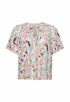 Vrouw draagt een losse veelkleurige blouse met korte mouwen en een strikje bij de hals, en zwarte broek tegen een oranje achtergrond.