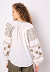 Weiße Bluse mit gestickten goldenen und braunen Mustern an den Ärmeln, mit elastischen Bündchen und einem gerafften Rückendesign. Glatte Textur, lässige Passform.
