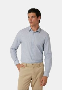 Camicia a righe blu chiaro con colletto a bottone, design aderente, maniche lunghe e tessuto di cotone liscio. Indossata con chinos color sabbia.