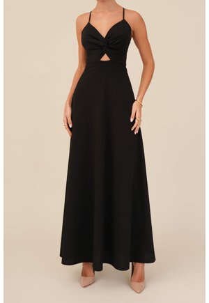 Woman wearing a sleeveless black maxi dress with twisted knot and cutout at the front, paired with beige pointed heels and gold jewelry.