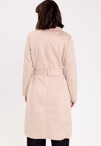 Cappotto lungo in suede beige con vita regolabile, dettagli nella cucitura posteriore e maniche lunghe. Texture liscia, design minimalista, silhouette di lunghezza intermedia.