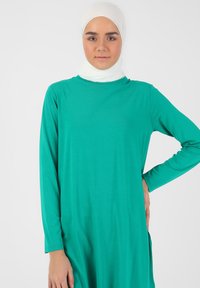 Turkis langærmet kjole med løs pasform og bådudskæring, parret med et hvidt tekstureret tørklæde. Simpelt design uden yderligere mønstre.
