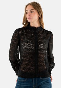 Blusa nera in pizzo con un motivo a diamante testurizzato, collo alto, maniche lunghe a sbuffo e chiusura frontale con bottoni. Abbinata a jeans blu.