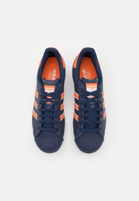 Námořnické modré tenisky s oranžovými akcenty a bílými pruhy. Disponují texturovanou gumovou špičkou a designem na šněrování. Obsahují značení "adidas".