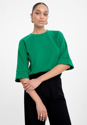 Green top with a round neckline, elbow-length sleeves, and a relaxed fit. Styled with black trousers and gold ring accents on the hands.