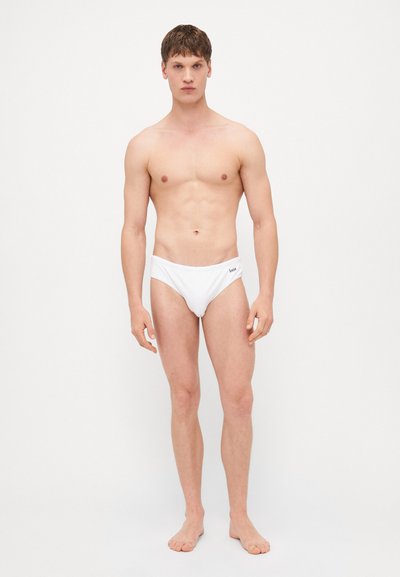 Slip de bain blanc avec une taille basse, fabriqué en tissu lisse, présentant un logo sur la ceinture, modèle debout pieds nus.