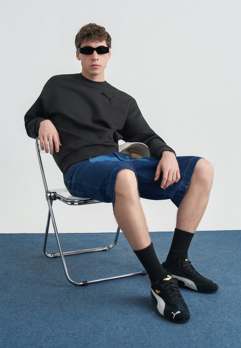 Νέος άνδρας που φοράει μαύρα γυαλιά ηλίου, μαύρο φούτερ, μπλε τζιν σορτς, μαύρα αθλητικά παπούτσια Puma με μαύρες κάλτσες, κάθεται σε μεταλλική καρέκλα πάνω σε μπλε χαλί.