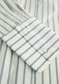 Cămașă din bumbac cu dungi verticale albe și bleumarin, având manșetă cu nasturi și o textură netedă și moale.