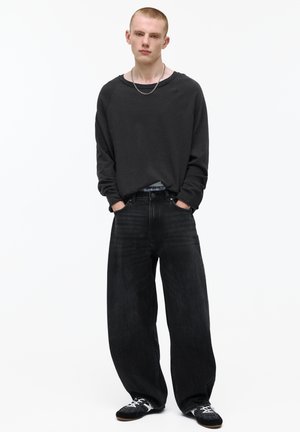 Giovane uomo in piedi con le mani nelle tasche, indossa jeans neri larghi, maglia nera a maniche lunghe, sneakers e una collana a catena d'argento.