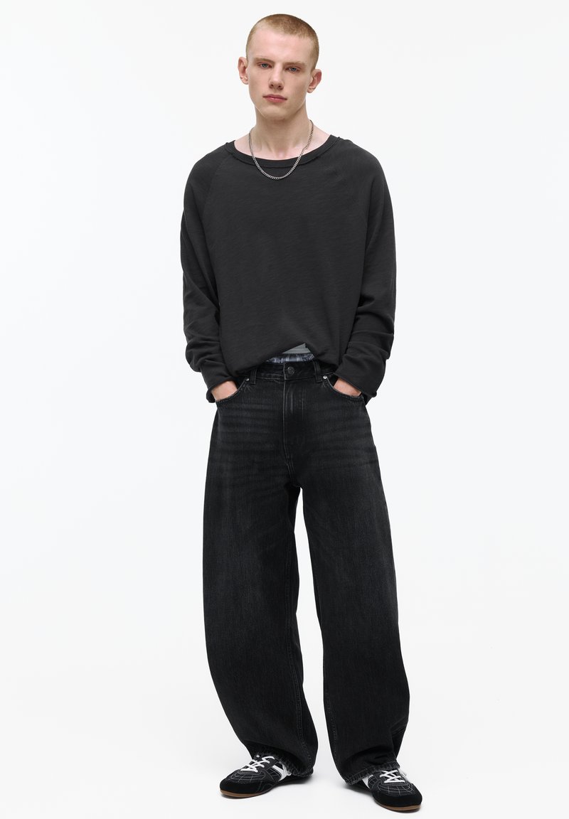 Junger Mann, der mit den Händen in den Taschen dasteht, trägt lockere schwarze Jeans, ein schwarzes Langarmshirt, Turnschuhe und eine silberne Kettenhalskette.