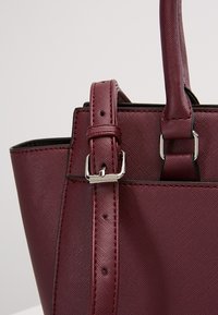 Gros plan d’un sac à main en cuir texturé rouge foncé montrant une sangle réglable avec boucle argentée et une poignée robuste attachée avec des anneaux métalliques.