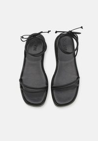 Sandali piatti neri realizzati in materiale liscio, con cinturini incrociati e un design con lacci, dotati di punta quadrata e un'estetica minimalista.