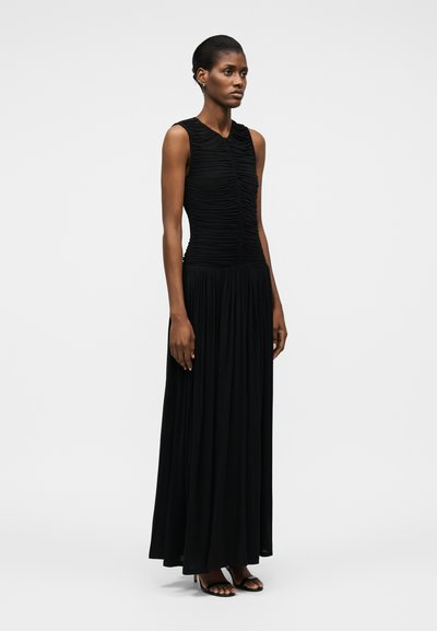 Naine, kes kannab musta varrukateta maksikleit ruššiitud büstiga ja volditud seelikuga, seisab käed lõdvalt kõrval, jalas mustad kontsakingad.