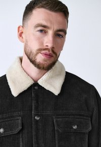 Chaqueta de pana negra con un cuello de borrego sintético color crema, con cierre de botones en la parte delantera, bolsillos en el pecho y tejido texturizado.
