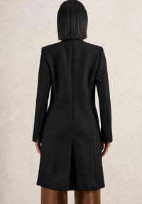 Schwarzer Wollmantel mit taillierter Silhouette, langen Ärmeln, gerader Schnitt und einem mittigen Rückenschlitz, vor einem neutralen Hintergrund präsentiert.