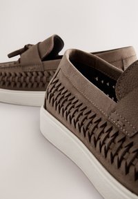 Brown leather slip-on shoes with woven side detailing, white rubber soles, and a smooth texture. A bow accent is present on the top.