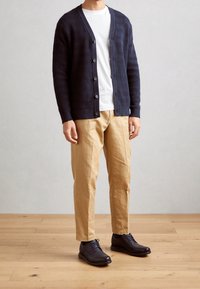 Homme portant un cardigan boutonné bleu marine, un t-shirt blanc, un pantalon beige et des chaussures habillées marron foncé, debout sur un sol en bois devant un mur uni.