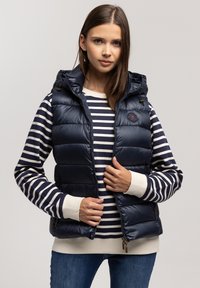 Granatowa kamizelka puchowa z kapturem, z zamkiem z przodu i bocznymi kieszeniami. Nosi się ją na granatowej i białej bluzce z długim rękawem w paski.