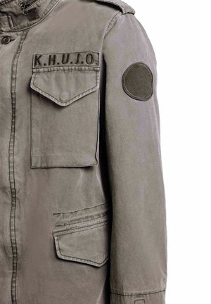 Grøn militærinspireret jakke med klaplommer foran, skulderrem, rund patch på ærmet og "K.H.U.J.O." broderet over brystlommen.