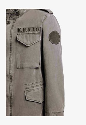 Veste de style militaire verte avec poches à rabat sur le devant, épaulette sur l'épaule, écusson circulaire sur la manche, et "K.H.U.J.O." brodé au-dessus de la poche poitrine.