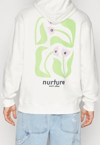 Sweat à capuche en coton blanc arborant un motif au dos avec des formes vertes, des fleurs roses, et le texte "prenez soin les uns des autres" et "amour".