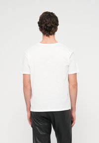 Λευκό βαμβακερό μπλου ζάκι με κοντά μανίκια, με στρογγυλή λαιμόκοψη και ίσιο τελείωμα. Φορεμένο με μαύρο παντελόνι, φαίνεται από πίσω.