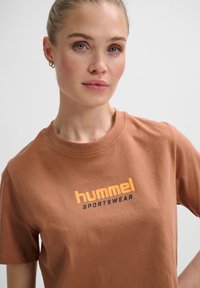 Brun bomulds T-shirt med rund halsudskæring og korte ærmer, med "hummel" logoet trykt i orange og sort på fronten.
