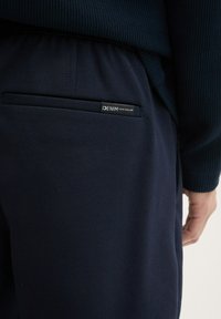 Granatowe spodnie z teksturowanej tkaniny z tylną kieszenią, na której znajduje się czarna metka z napisem "DENIM TOM TAILOR."