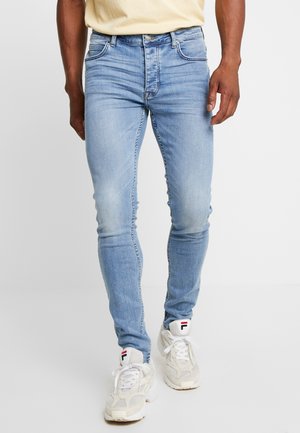 Man draagt lichtblauwe slim-fit jeans en witte sneakers met een logo op de tong, staand tegen een effen achtergrond.
