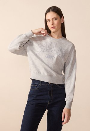 Grijze cropped sweatshirt met "DALLAS 1967" tekst, geribbelde manchetten en zoom, gecombineerd met donkere denim jeans. Zacht katoen, relaxte pasvorm.