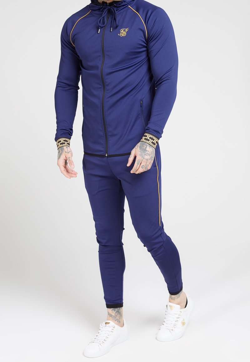 Navy athletic tracksuit with gold accents, featuring a full zip jacket with a hood, tapered pants, and contrasting side stripes.