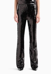 Pantalones de lentejuelas negras con piernas acampanadas, que presentan una superficie brillante y texturizada que refleja la luz. Diseño ajustado, combinados con zapatos negros.