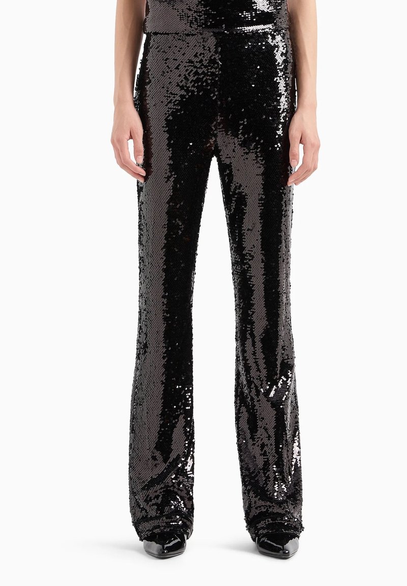 Pantalones de lentejuelas negras con piernas acampanadas, que presentan una superficie brillante y texturizada que refleja la luz. Diseño ajustado, combinados con zapatos negros.