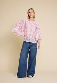 Lys rosa bluse med blomsterdesign og vide, plisserte ermer, parret med løse, rett ben jeans i blått og hvite sneakers. Nøytral bakgrunn.