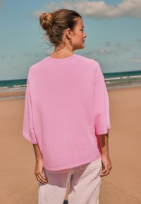 Top roz, lejer, cu mâneci scurte, realizat dintr-un material moale, asortat cu pantaloni albi cu textură. Fundalul prezintă o plajă și o vedere spre ocean.