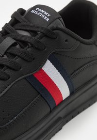 Czarne trampki z teksturowaną skórzaną cholewką, płaskimi sznurowadłami i bocznym paskiem w kolorze czerwonym, białym i granatowym. Logo na języku z napisem „Tommy Hilfiger”.}}