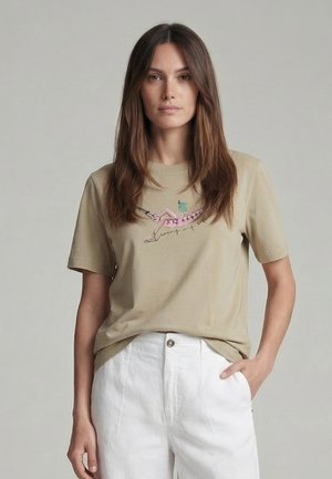 Woman with long brown hair wears beige t-shirt with pink flamingo graphic and white high-waist pants, standing against plain gray background.