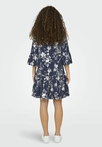 Robe bleu marine à motifs floraux, avec des manches cloche et un ourlet à volants superposés. Fabriquée en tissu léger, associée à des baskets blanches.