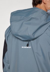 Blue lightweight jacket with a hood, featuring a textured surface and black accents. "MAMMUT" logo in white on the back. Drawstring detail at the hood.
