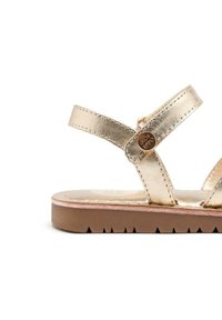 Sandal in metallo dorato con due cinghie incrociate e un accento decorativo in metallo. La suola è marrone chiaro con bordo seghettato per una migliore aderenza.