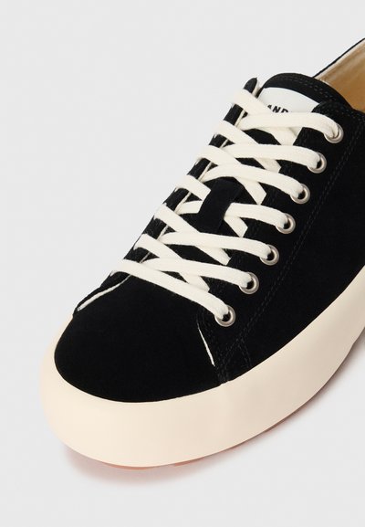 Must must-sneaker valge pingutusnööriga, ümmarguse varba ja kreemja kummitald ega. Omab hõbedasi silmade ja väikest logo keelel.