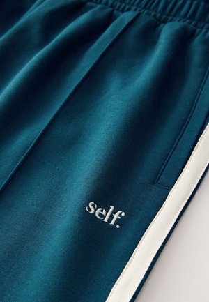 Teal mjukisbyxor med mjuk textur, med en vit sidrand och broderad "self."-logotyp i ljus färg nära fickområdet.