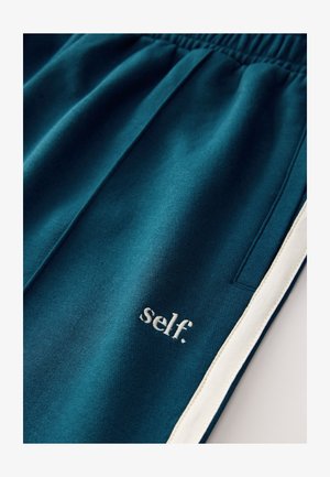 Tirkizne trenirka s mekanom teksturom, s bijelim bočnim prugama i izvezenim logotipom "self." svijetle boje blizu džepa.