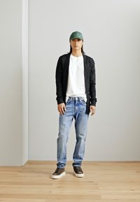 Casual outfit met een donkere cardigan, wit t-shirt, lichtblauwe jeans en bruine sneakers met witte zolen. Groene pet achterstevoren gedragen.