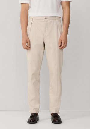 Mann trägt beigefarbene, maßgeschneiderte Hosen mit vorderen Falten, kombiniert mit dunkelbraunen Tassel-Loafers und einem weißen Kurzarmhemd, stehend.