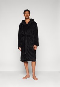 Black plush bathrobe with a hood, tie belt, and two front pockets. Soft texture and knee-length design. Model stands barefoot on a plain background.