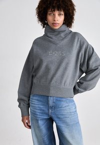 Grauer, taillierter Sweatshirt mit hohem Kragen und gerippten Bündchen; mit schimmerndem "BOSS"-Logo versehen. Kombiniert mit hellblauer Jeans.