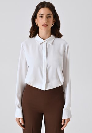 Blusa bianca a maniche lunghe con colletto classico, texture liscia e chiusura con bottoni. Abbinata a pantaloni marrone scuro.