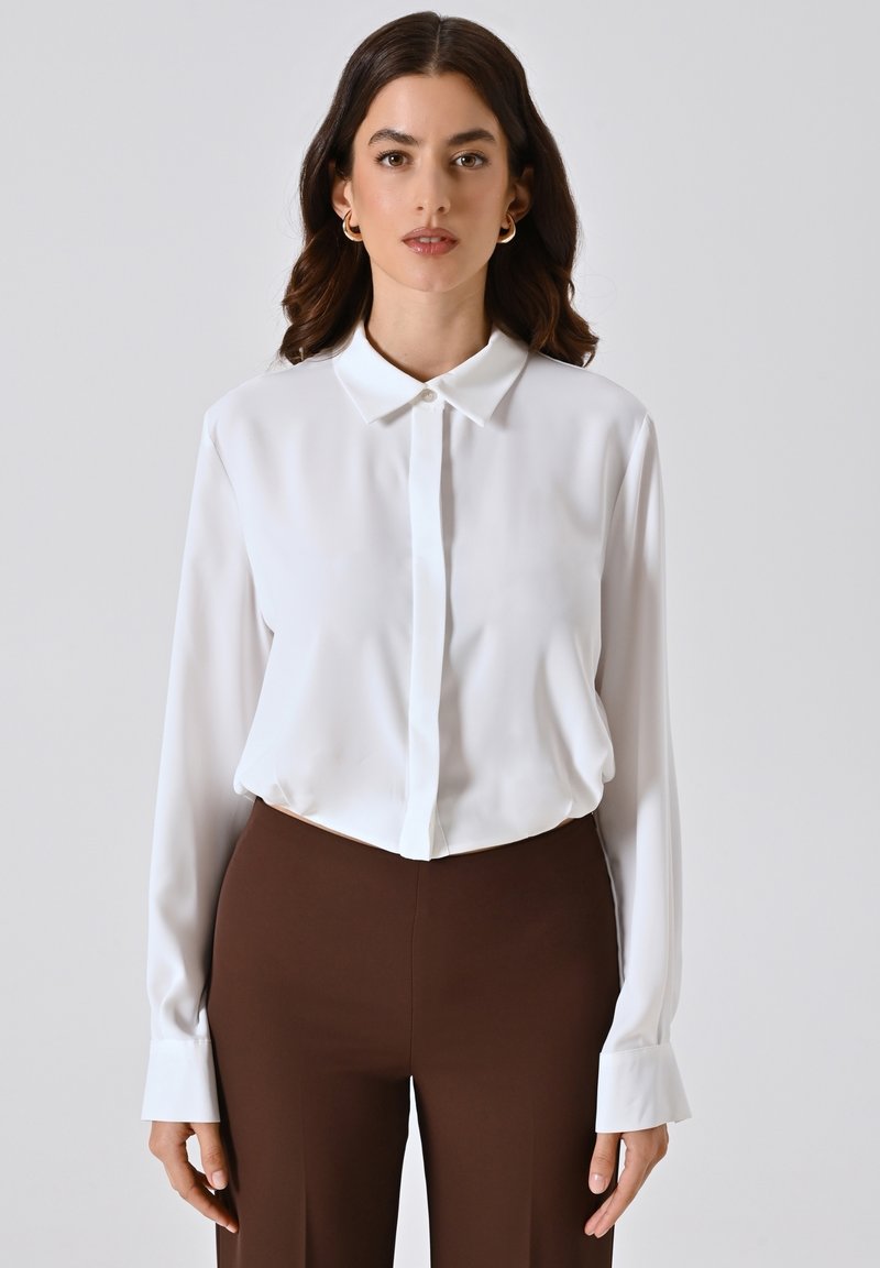 Blusa bianca a maniche lunghe con colletto classico, texture liscia e chiusura con bottoni. Abbinata a pantaloni marrone scuro.