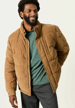 Mann lächelt und trägt eine braune gesteppte Jacke über einem blau-grün gestreiften Hemd mit dunkler Hose vor hellem Hintergrund.
